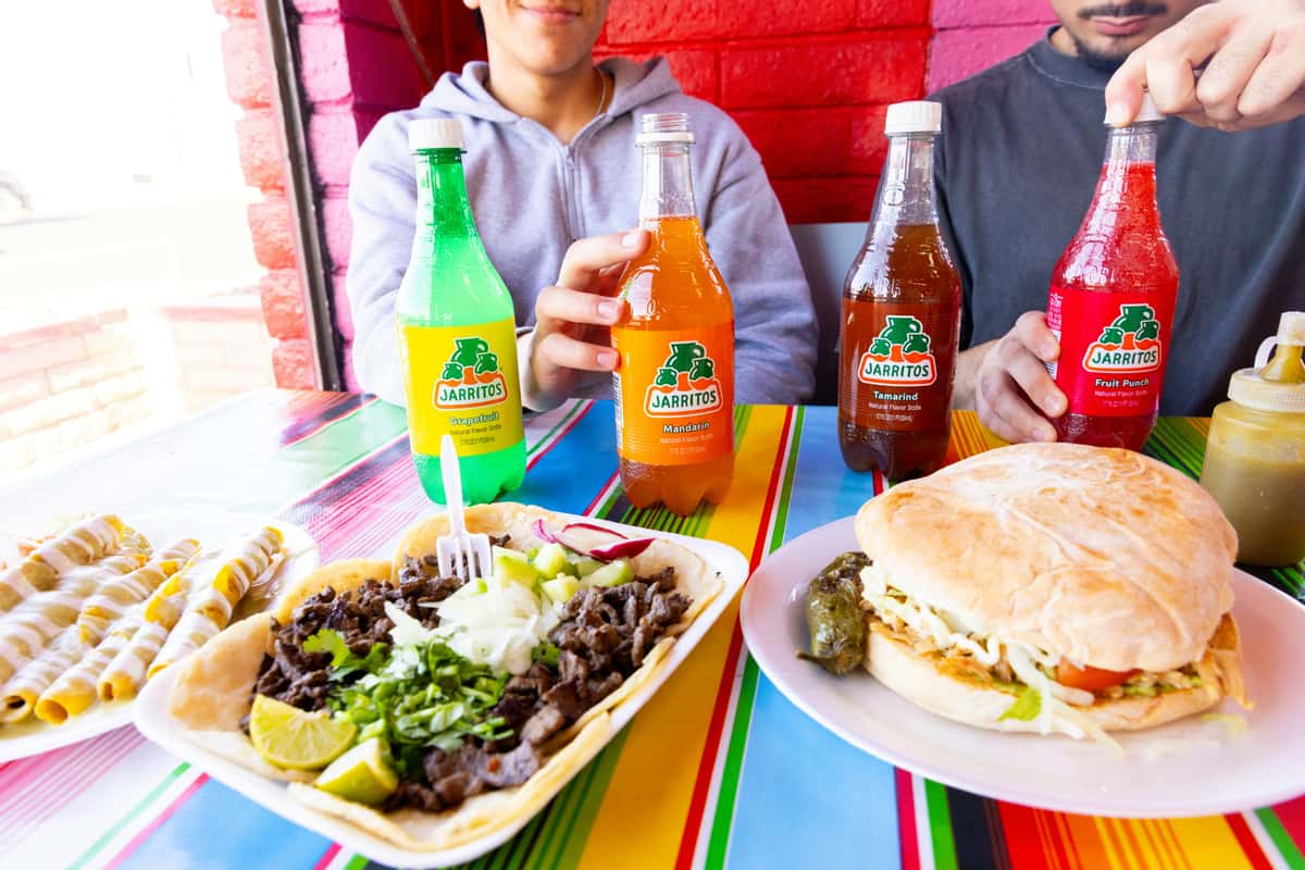 Mexican food spread with tacos, torta sandwich, taquitos, and assorted Jarritos sodas—vibrant casual Mexican meal.