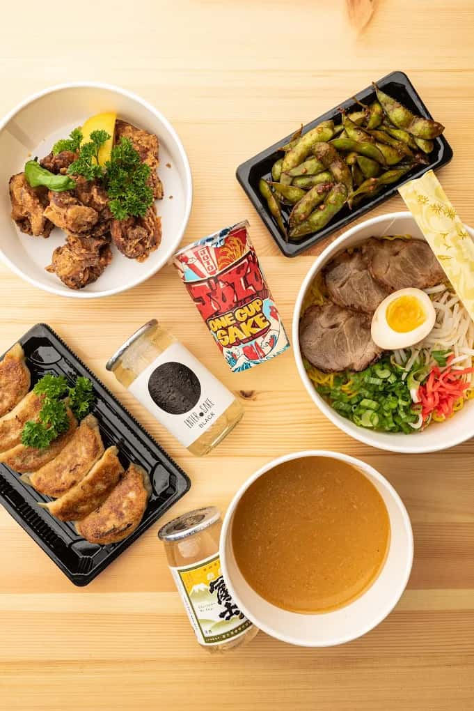 Mixed Japanese takeaway: fried chicken, edamame, gyoza, ramen, and a side of broth, with two sake bottles.