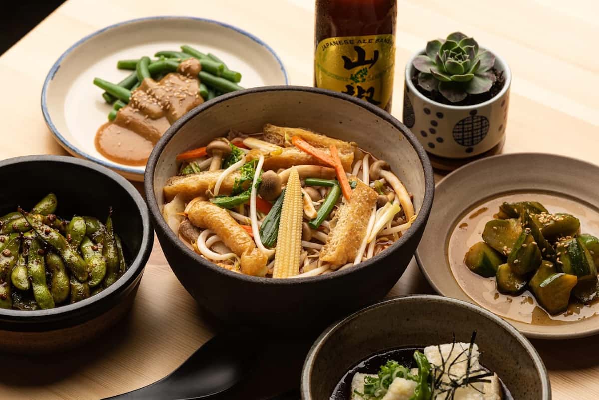 Udon noodle bowl with fried tofu, baby corn, and vegetables, surrounded by side dishes including edamame and green beans.