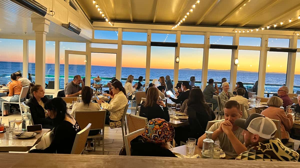 Busy evening crowd at Driftwood Kitchen enjoying dinner with panoramic ocean views through floor-to-ceiling windows, as the sun sets over the Pacific, casting a warm orange and blue glow.