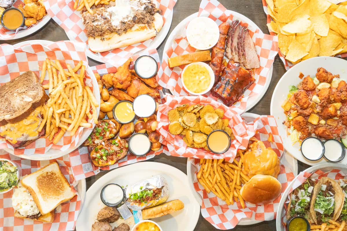 Overhead spread of bar food including BBQ ribs, brisket, wings, burgers, sandwiches, fried pickles, and fries on checkered paper-lined plates