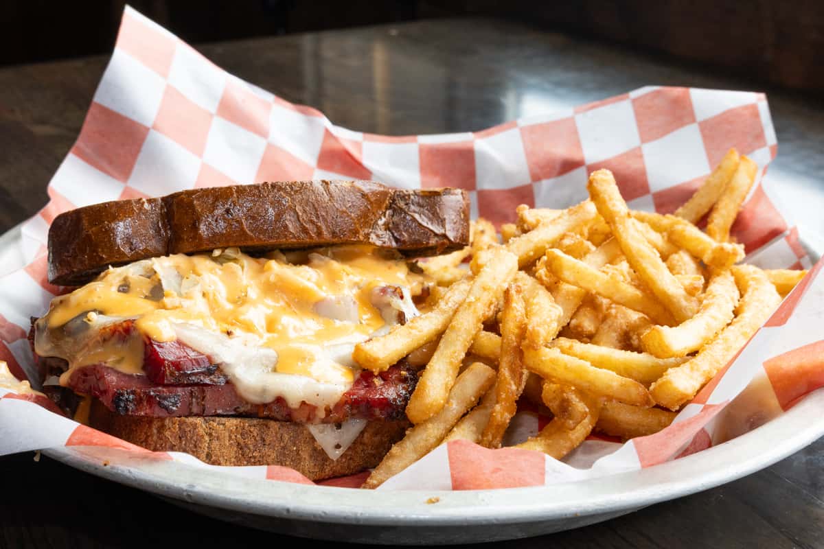 Grilled pastrami sandwich with melted Swiss and cheddar cheese on toasted rye, served with crispy French fries on a checkered deli tray