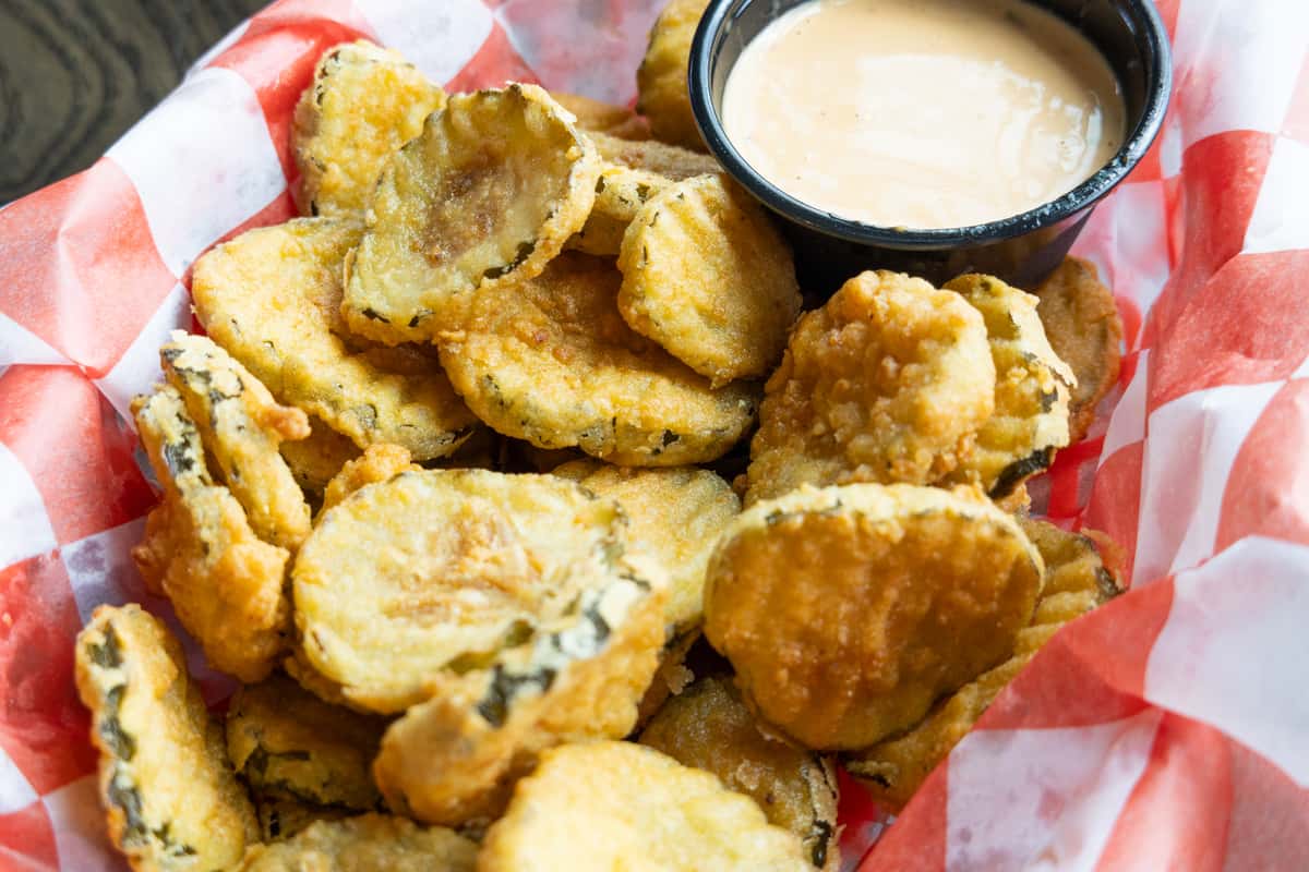 Crispy fried pickle chips served with creamy dipping sauce in a red checkered basket
