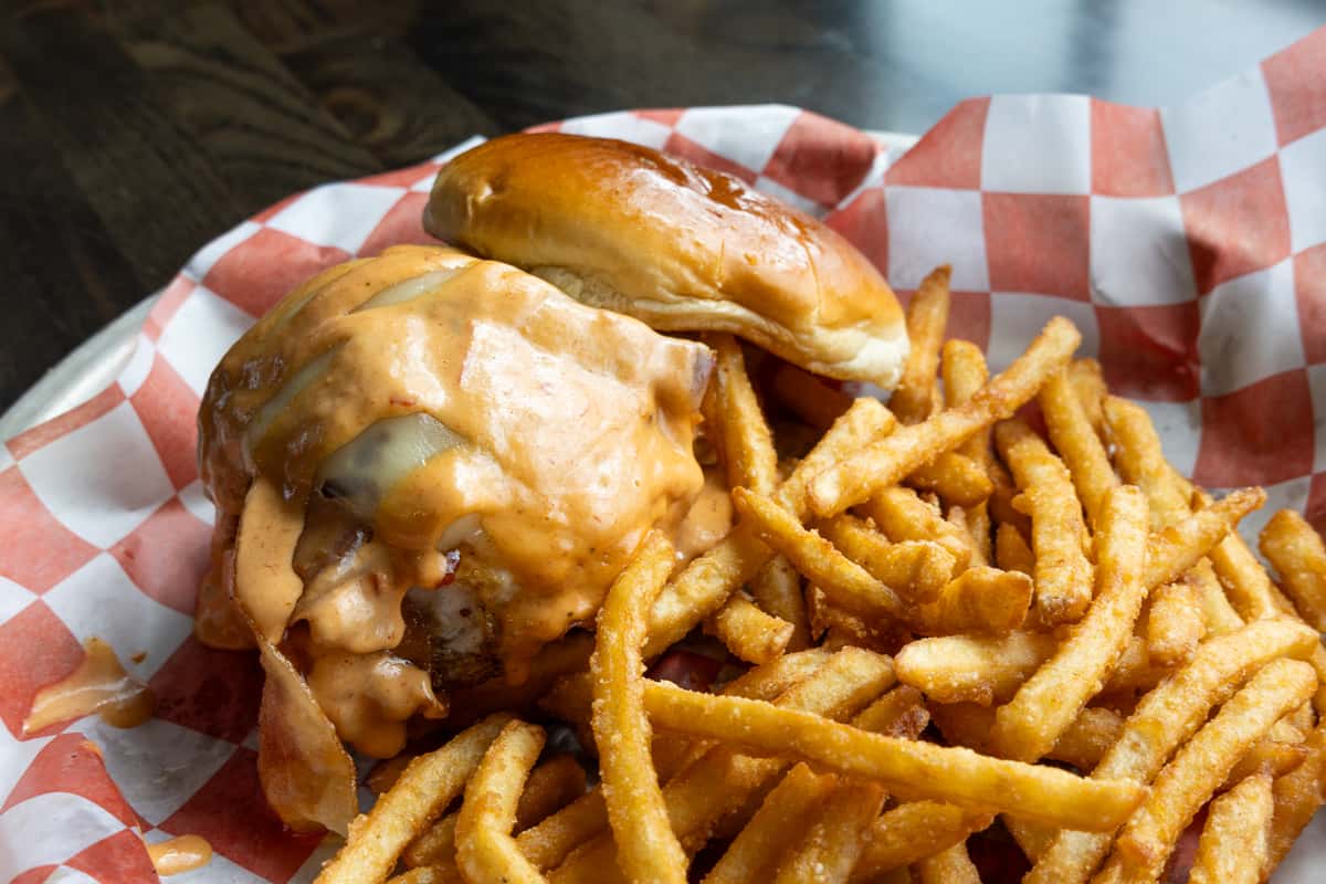 Juicy cheeseburger topped with melted cheese and creamy sauce on a toasted bun, served with crispy French fries on a checkered paper-lined plate