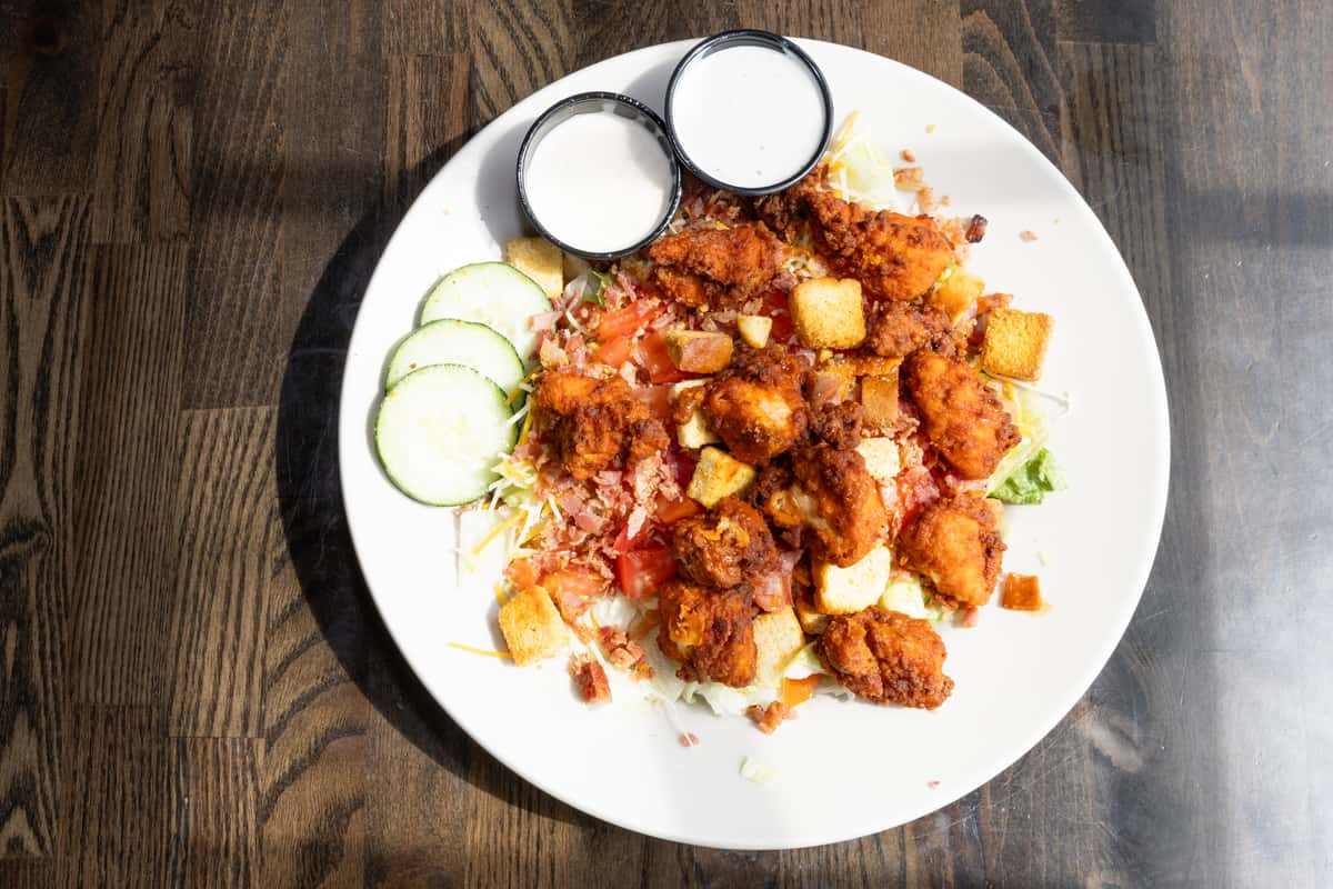 Overhead view of buffalo chicken salad with crispy fried chicken, bacon, tomatoes, cucumbers, shredded cheese, and ranch dressing on a white plate