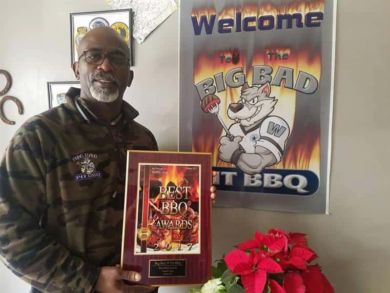Man holding Best BBQ Award 2017 in front of Welcome to the Big Bad Pit BBQ on wall.