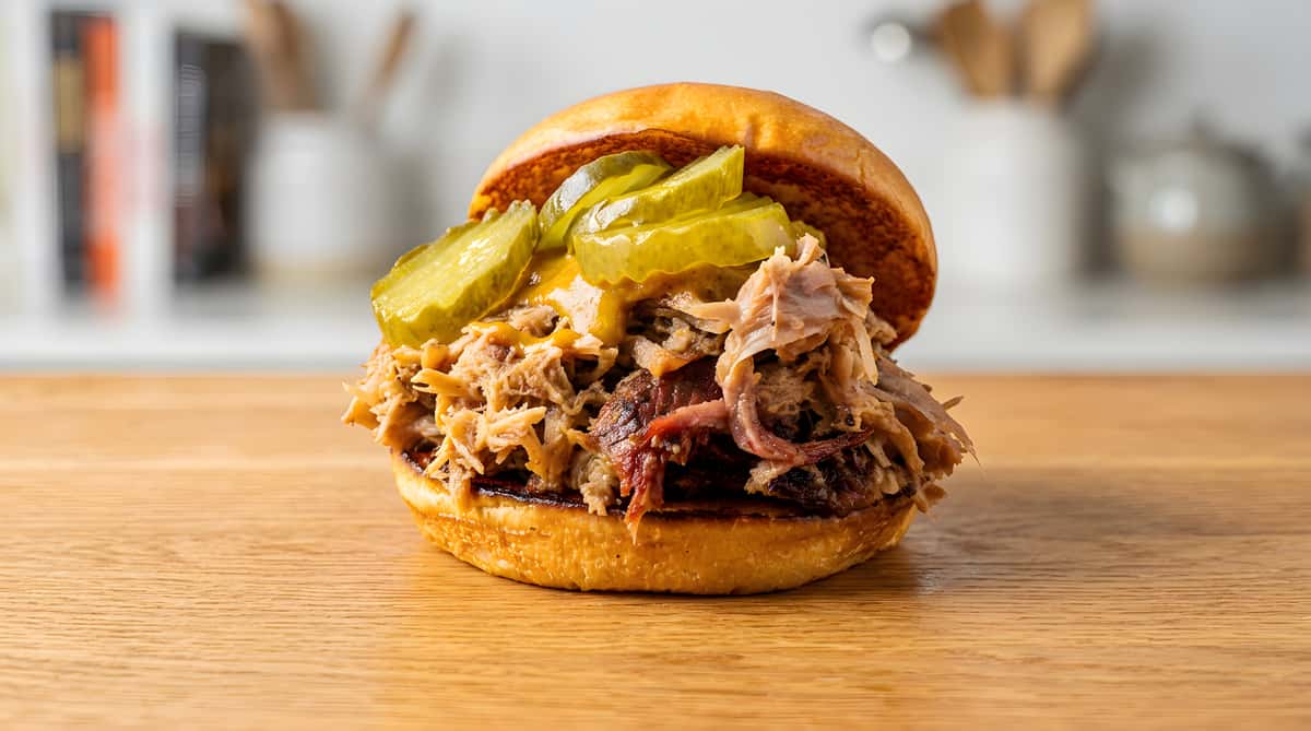 Pulled pork sandwich on a bun with pickles and mustard, on a wooden board.