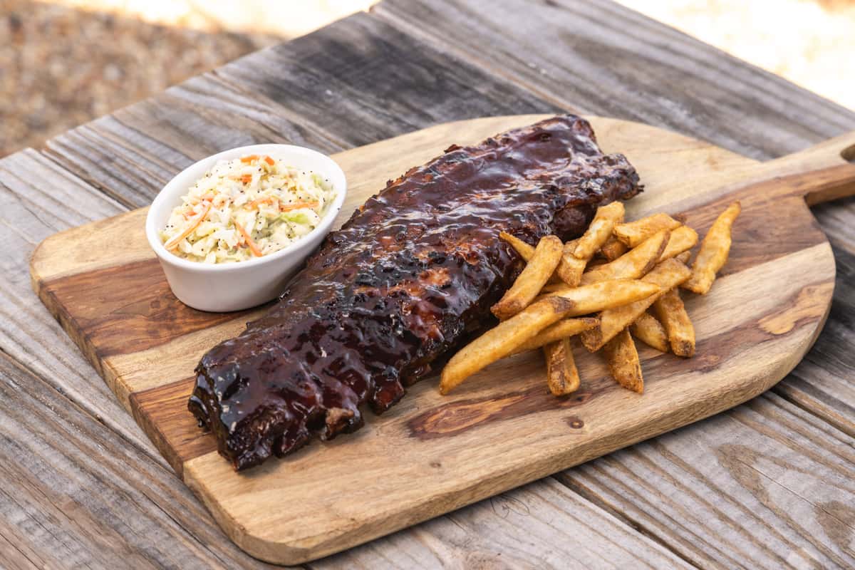 A slab of bbq ribs with a side of coleslaw and fries on a wooden platter