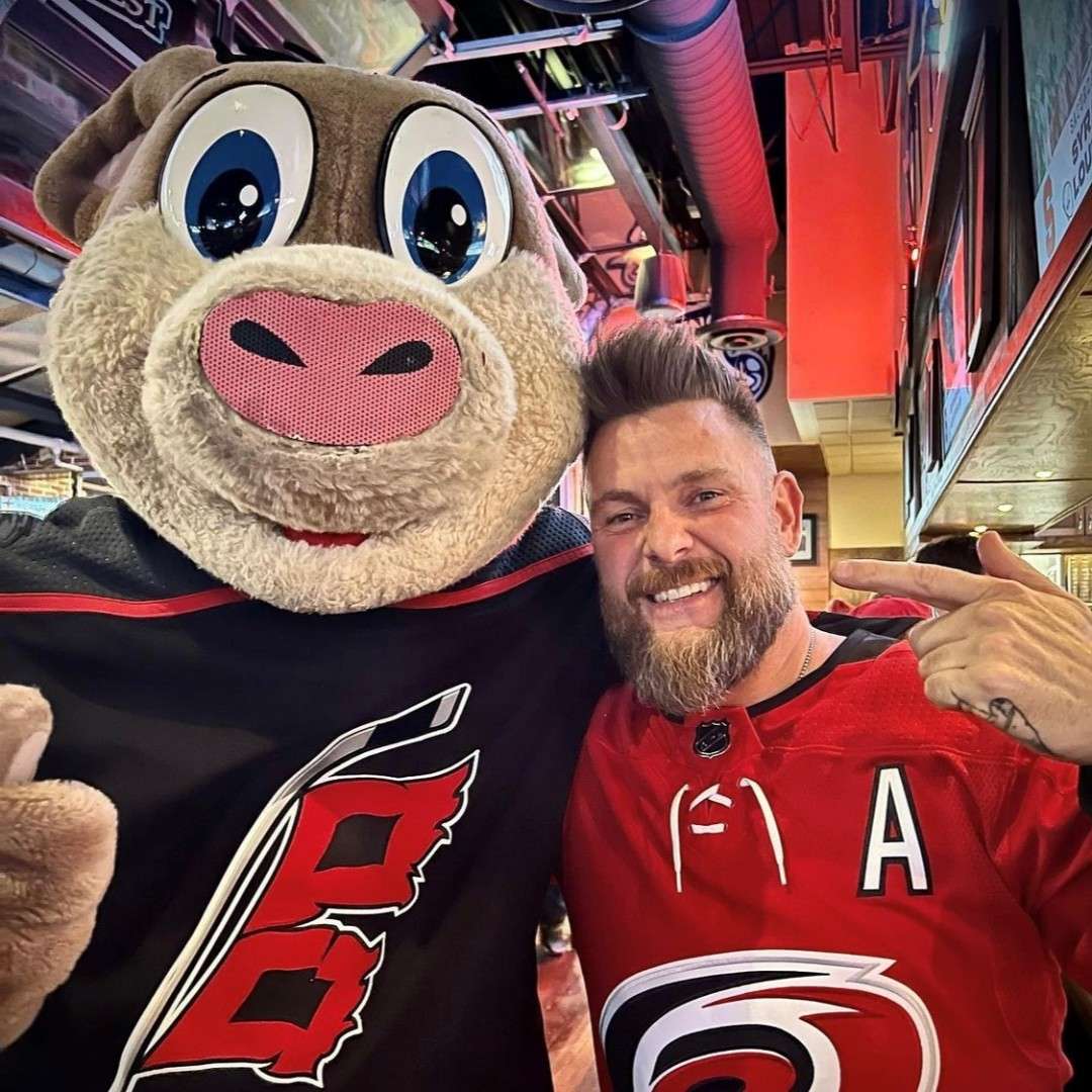 A smiling man in a Carolina Hurricanes jersey poses with Stormy the mascot at Carolina Ale House.
