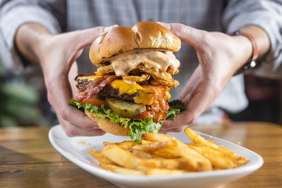 Hands holding a stacked cheeseburger in front of a plate of french fries