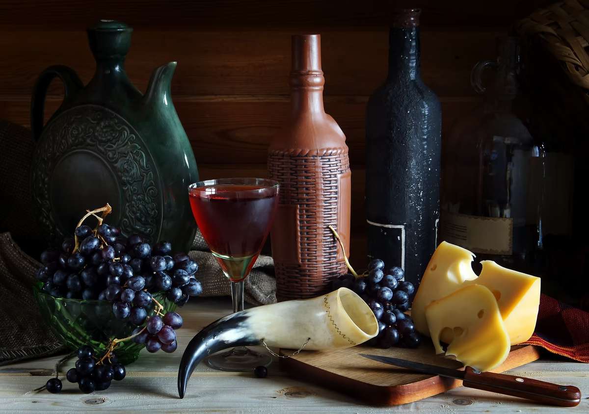 Rustic still life with wine bottles, a glass of red wine, cheese wedges, grapes, a drinking horn, and a decorative jug.