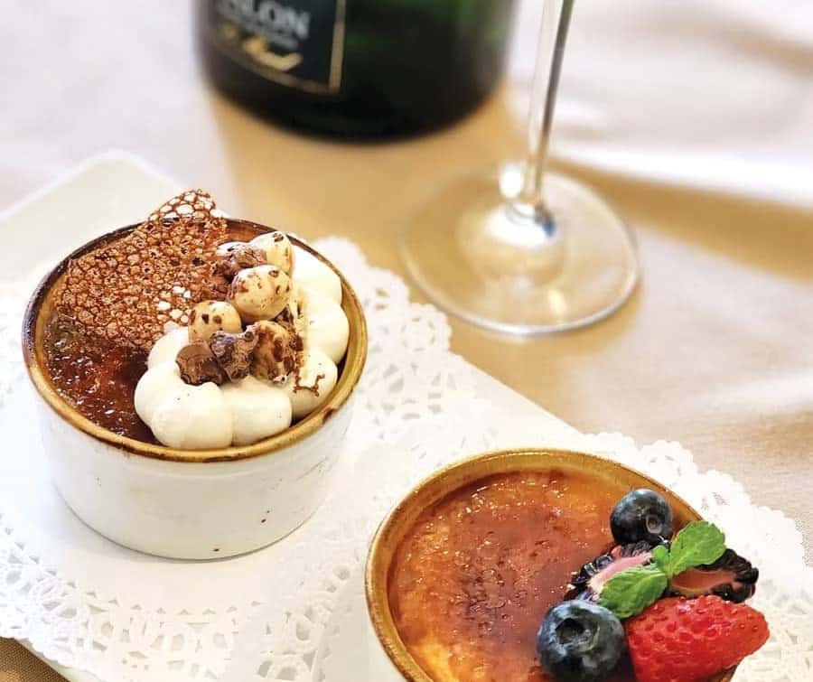 Two crème brûlées, one topped with berries and mint, the other with whipped cream, chocolate, and a tuile, beside a wine glass.