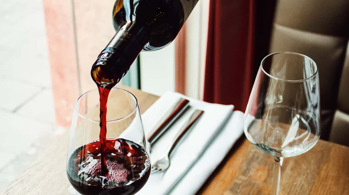 Red wine being poured into a glass on a wooden table set with silverware and a second empty wine glass.