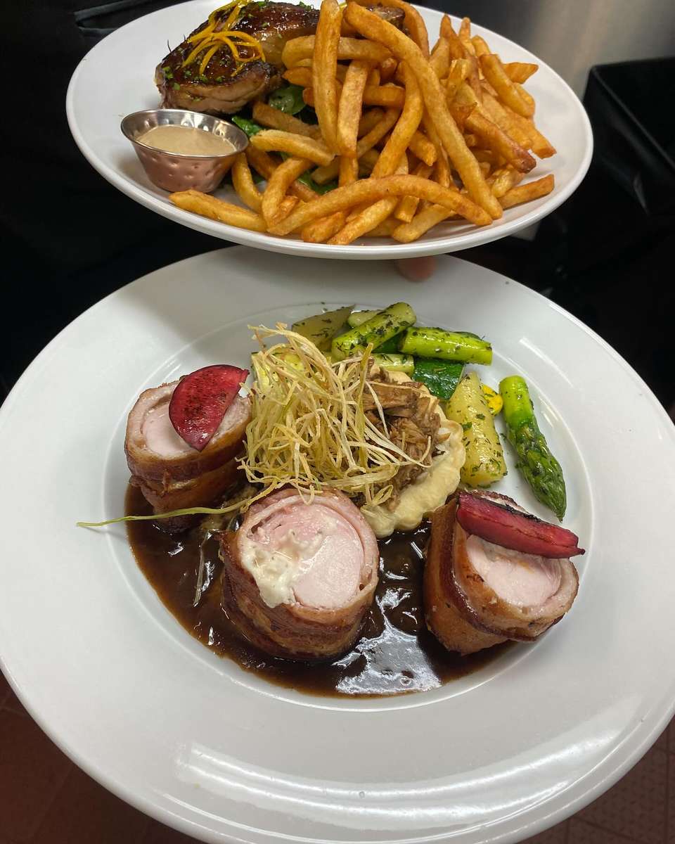 Bacon-wrapped chicken roulade with mashed potatoes, asparagus, and demi-glace; background plate with steak frites and sauce.
