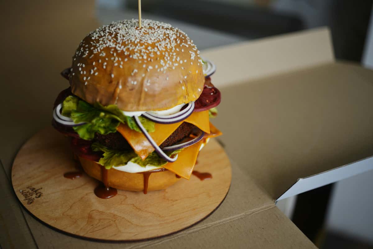 Giant gourmet burger cake designed to look like a cheeseburger with layers of fondant lettuce, cheese, onions, and tomato on a sesame seed bun, presented on a wooden cake board.
