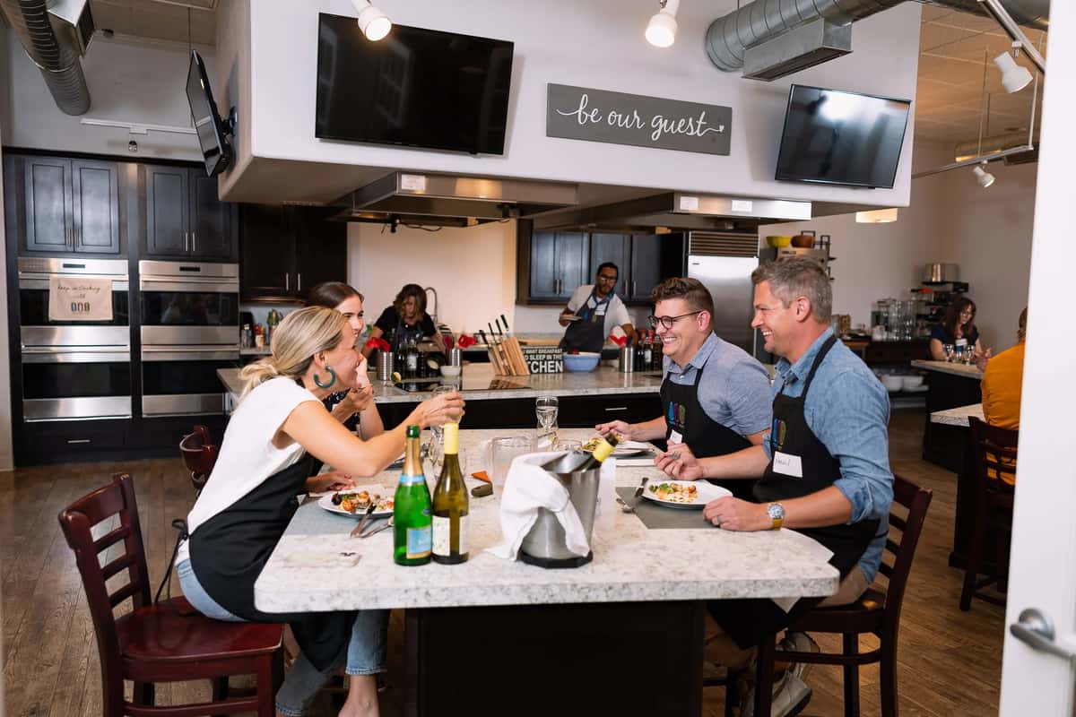 Guests enjoying a hands-on cooking class at a modern culinary studio with wine, shared dining table, and open kitchen atmosphere