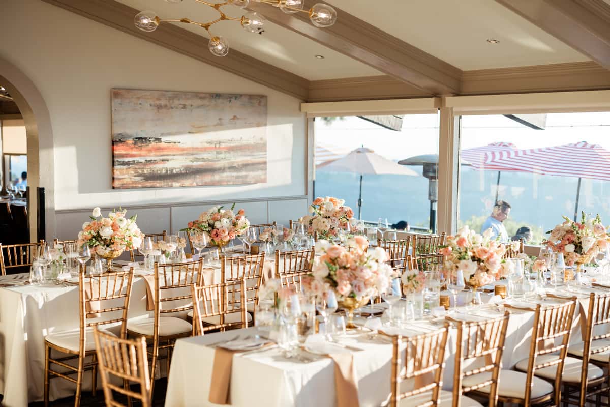 Elegant wedding reception dining room with floral centerpieces, gold chiavari chairs, and panoramic ocean-view windows in an upscale event venue