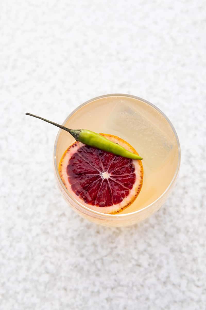 Top view of a yellow cocktail with a blood orange and chili garnish sitting on a marble counter top