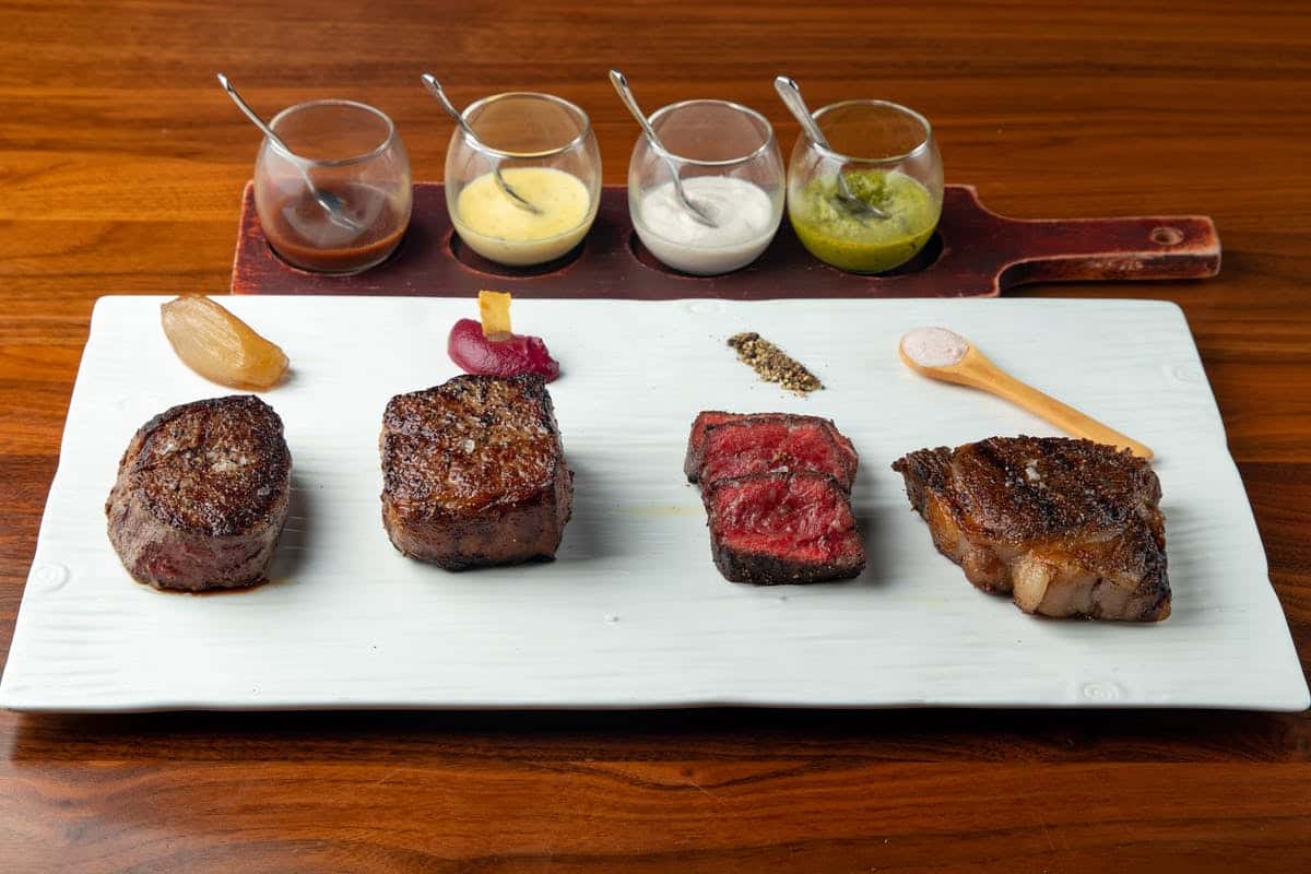 Four exquisite steaks artfully arranged on a white plate, accompanied by a quartet of flavorful sauces.