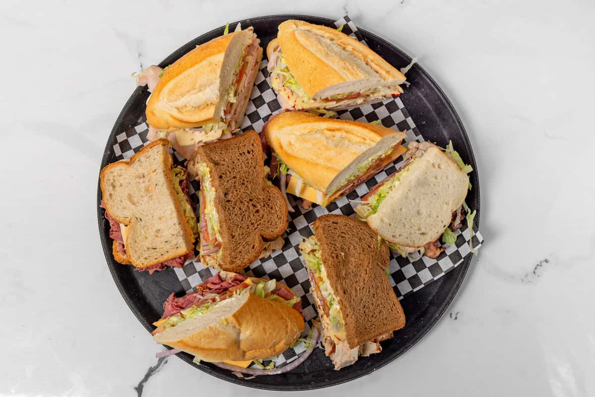 Assorted deli sandwich halves with sliced meats, cheese, lettuce, and tomatoes on mixed breads served on a tray