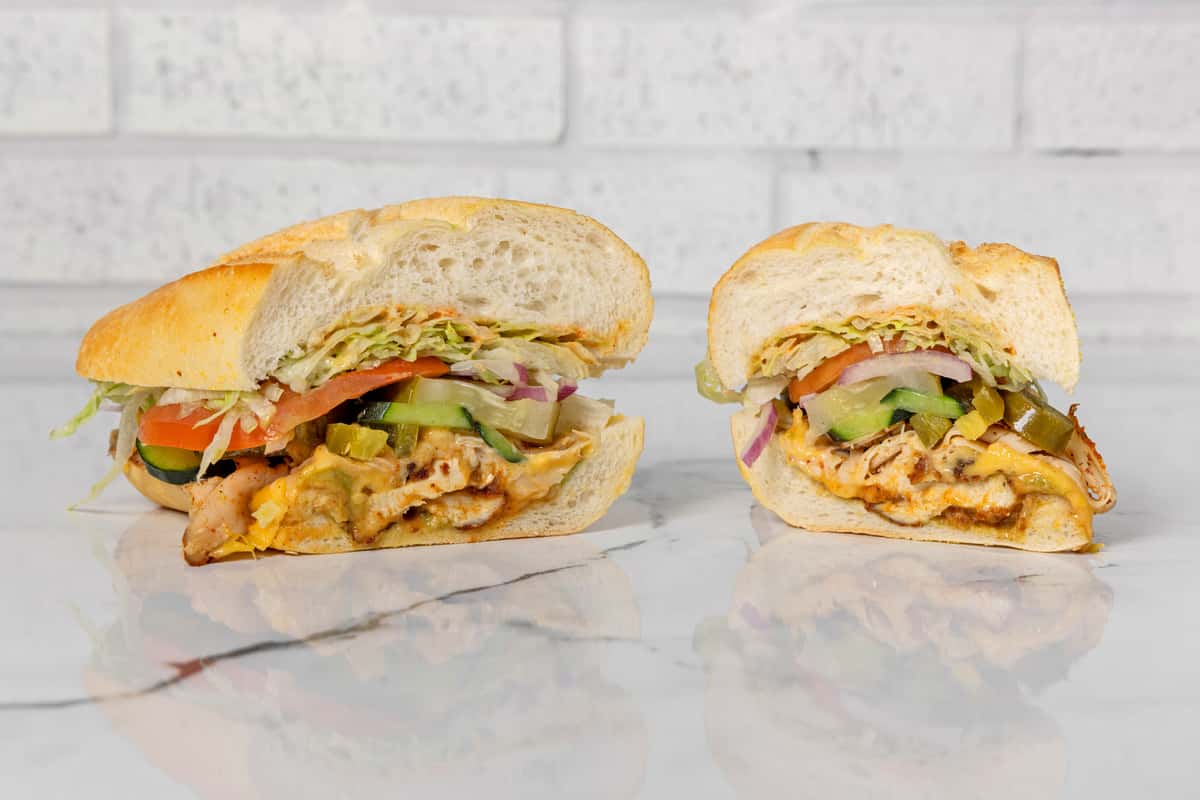 Grilled chicken sandwich halves with melted cheese, lettuce, tomato, pickles, and onions on a toasted hoagie roll