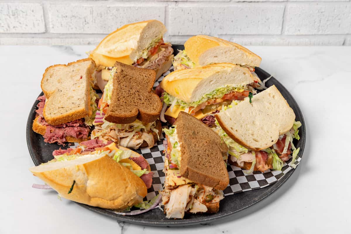 Assorted deli sandwich platter with turkey, pastrami, and cheese on white and whole grain bread, topped with lettuce, tomato, onions, and pickles on a serving tray