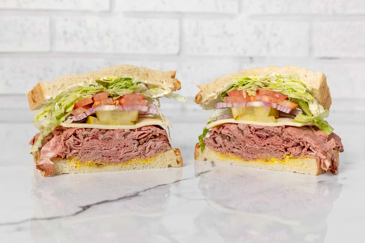 Pastrami sandwich halves with melted cheese, lettuce, tomato, red onion, and pickles on white bread, stacked deli-style on a marble surface