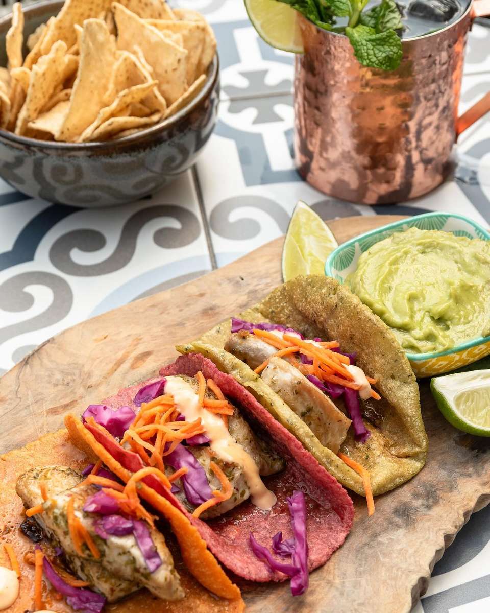 Trio of grilled fish tacos served on vibrant orange, pink, and green corn tortillas, topped with shredded purple cabbage, carrots, and creamy chipotle sauce, accompanied by fresh guacamole, lime wedges, crispy tortilla chips, and a copper mug cocktail—ideal for Baja coastal cuisine and taco lovers.