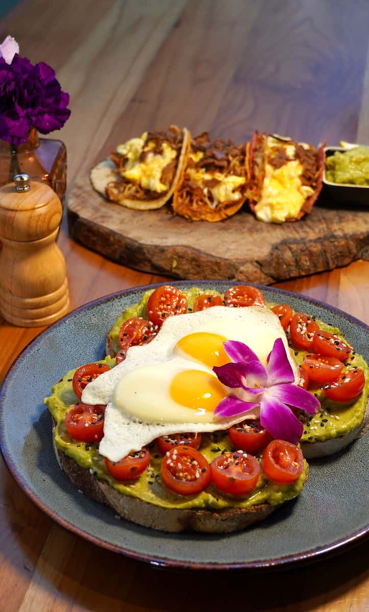 Avocado toast topped with cherry tomatoes, sunny-side-up eggs, and a purple orchid, with a background plate of tacos filled with scrambled eggs and meat.