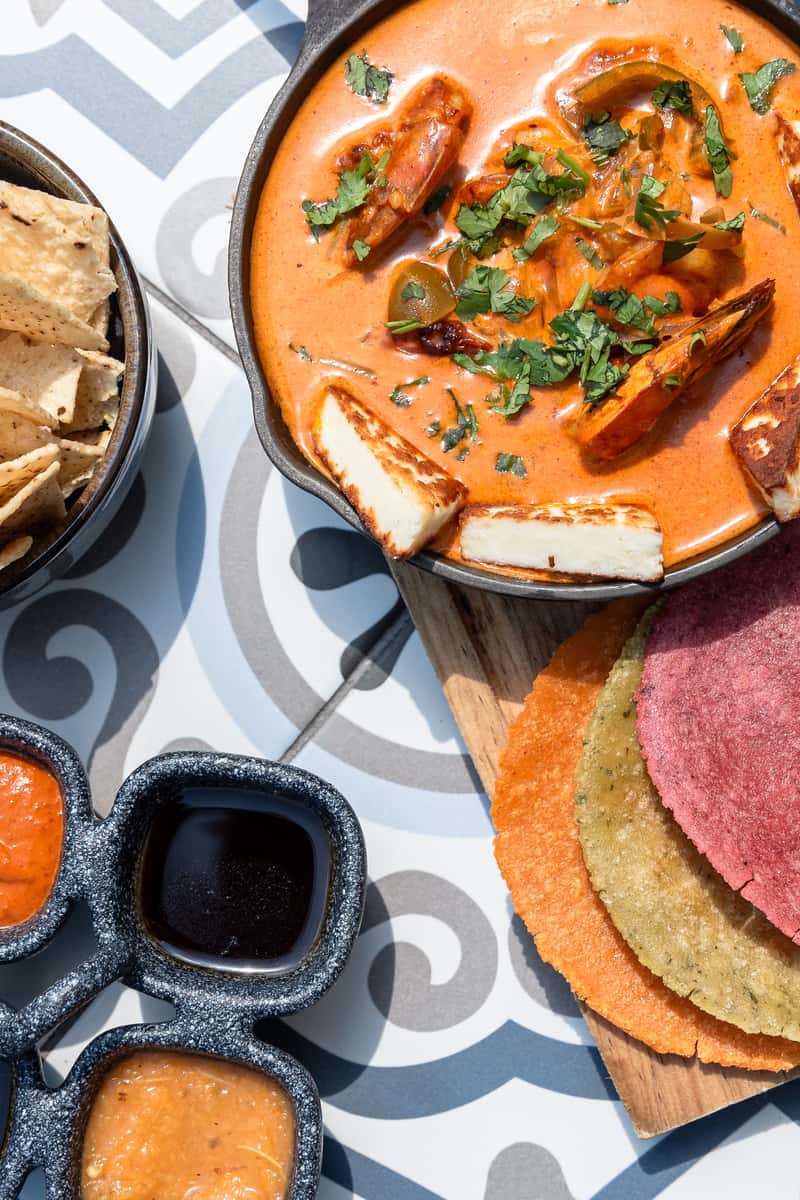 Creamy shrimp stew topped with fresh cilantro and grilled halloumi, served with colorful corn tortillas, house salsas, and tortilla chips—perfect for showcasing modern coastal Mexican cuisine and bold flavors.