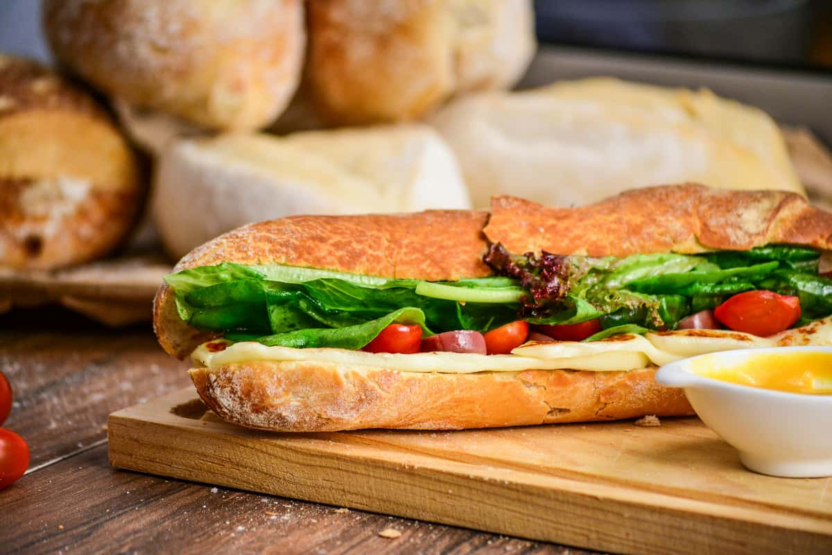 Fresh baguette sandwich with melted cheese, crisp lettuce, cherry tomatoes, and olive oil on a wooden cutting board.