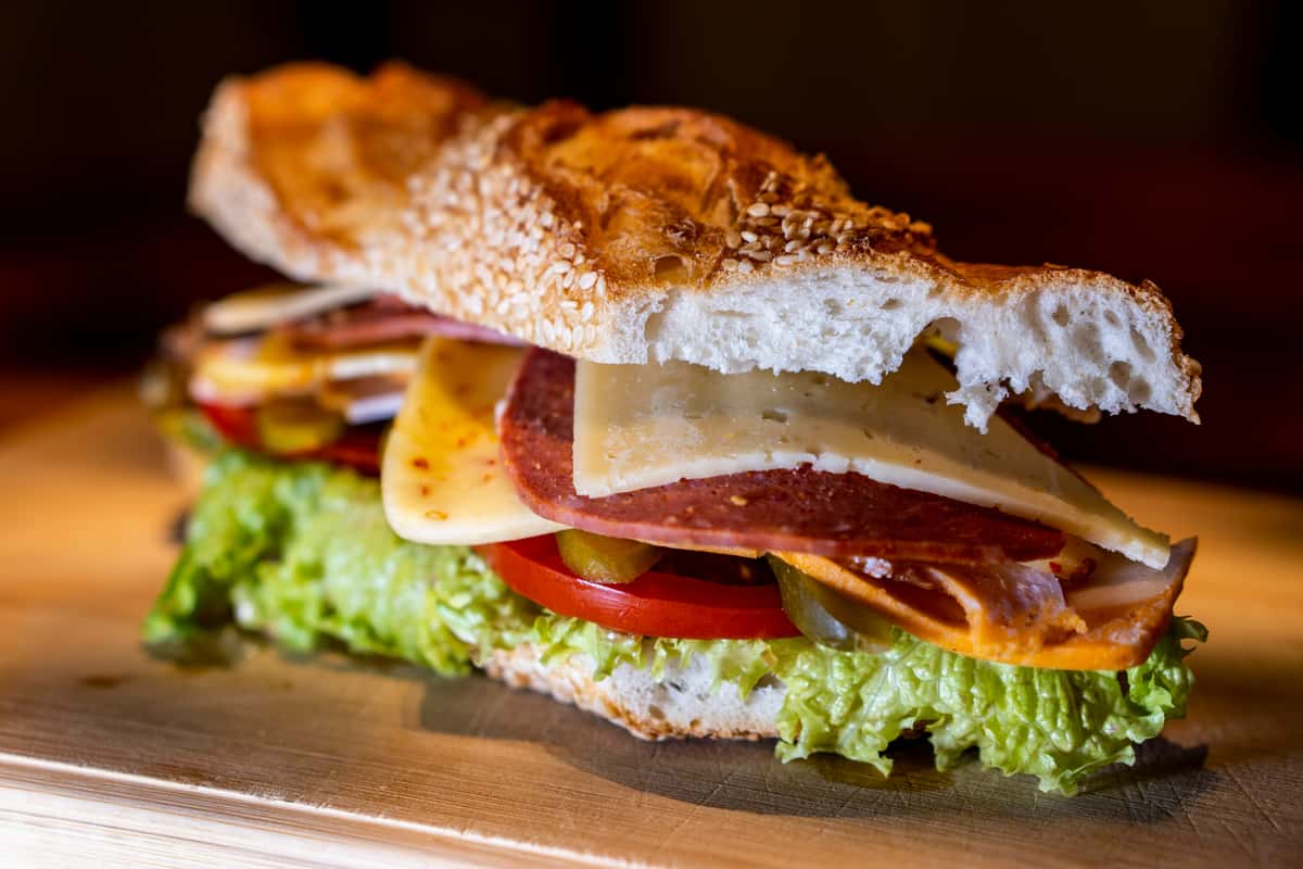 Italian sub sandwich with salami, sliced cheese, pickles, tomatoes, and lettuce on sesame-crusted bread.