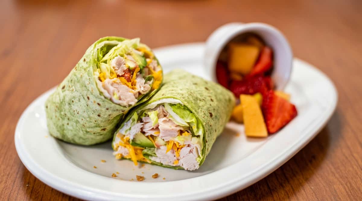 Two halves of a spinach wrap with turkey, cheese, lettuce, and avocado, served with a side of fruit.