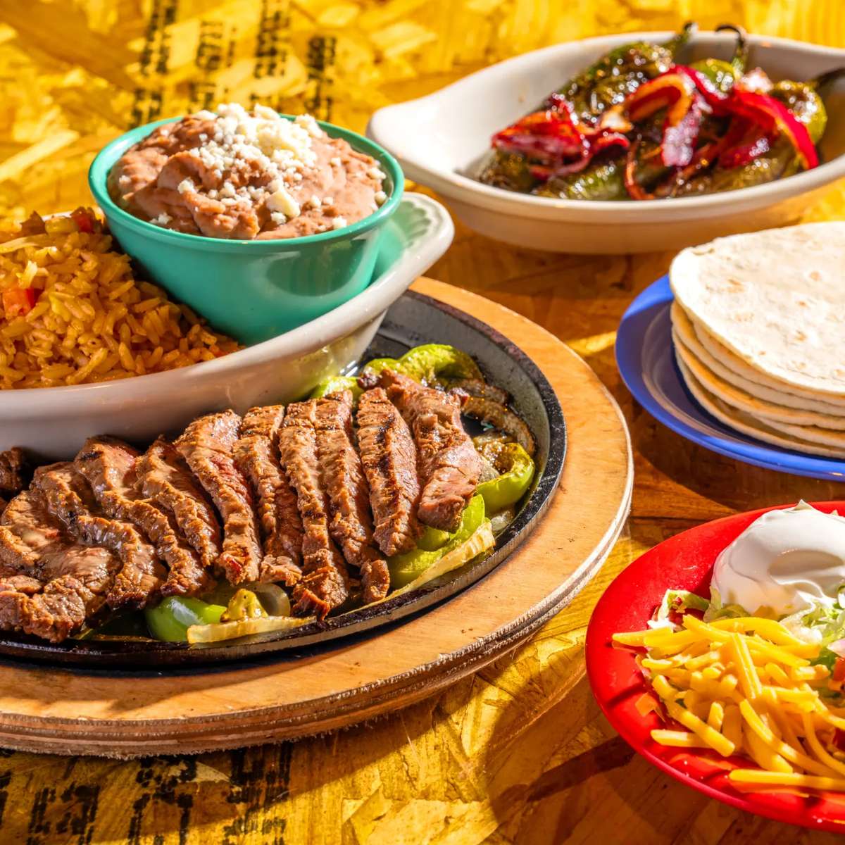 Sizzling steak fajitas with rice, refried beans, tortillas, and grilled peppers on a colorful Mexican table setting