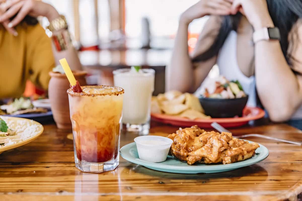 Chili-rimmed cocktail and boneless wings with dipping sauce on table, friends dining in casual restaurant setting