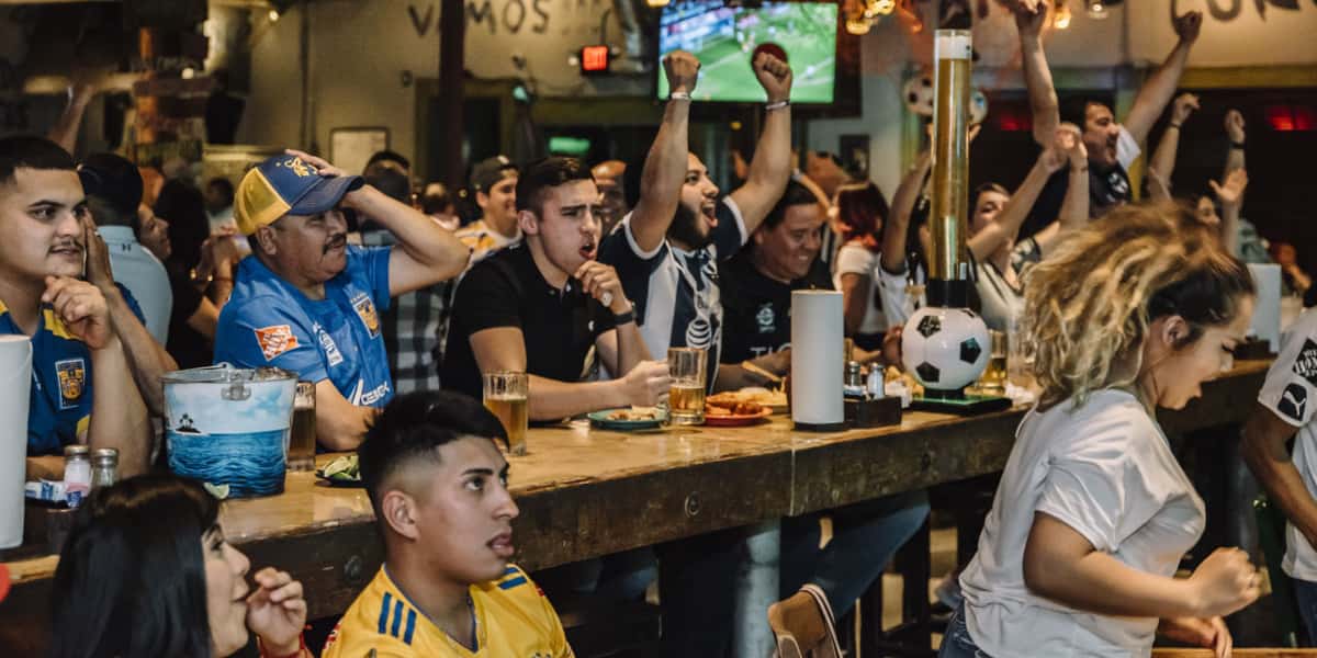Crowd of soccer fans cheering at sports bar, raising arms as game plays on big screens above bar tables