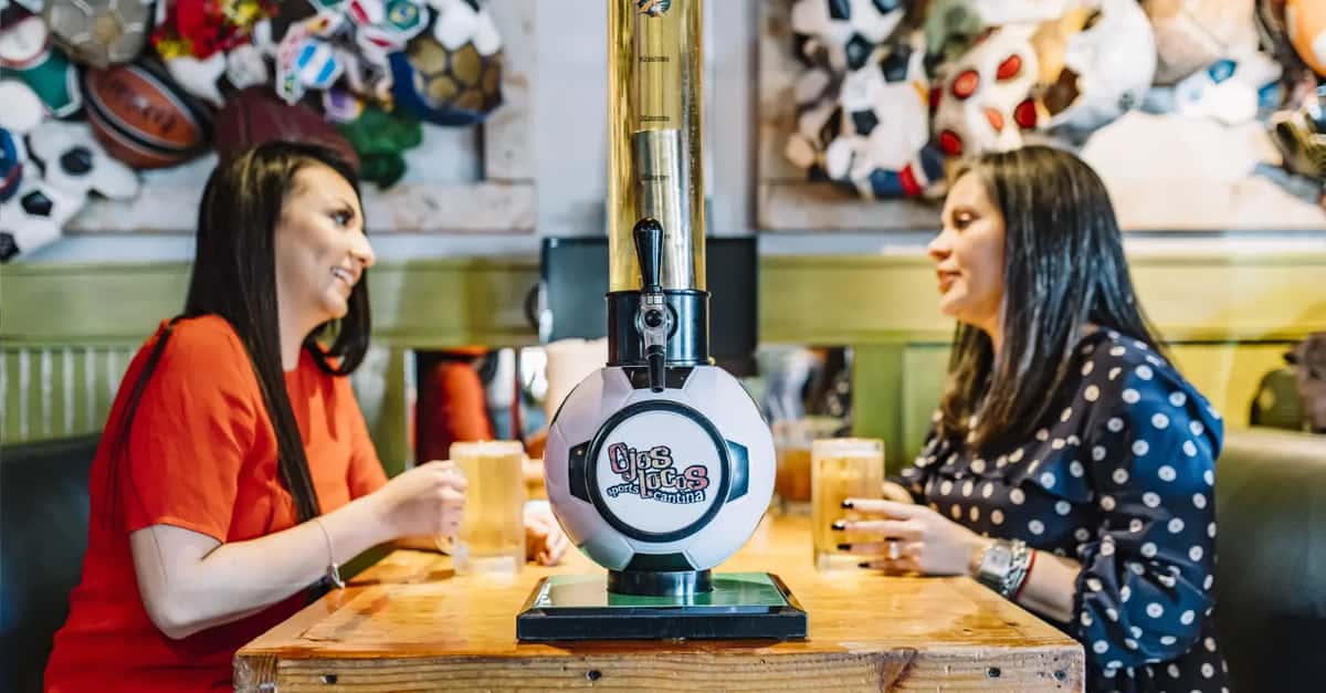 Two women enjoying beers at a table with Ojos Locos sports cantina beer tower between them, sports decor on wall