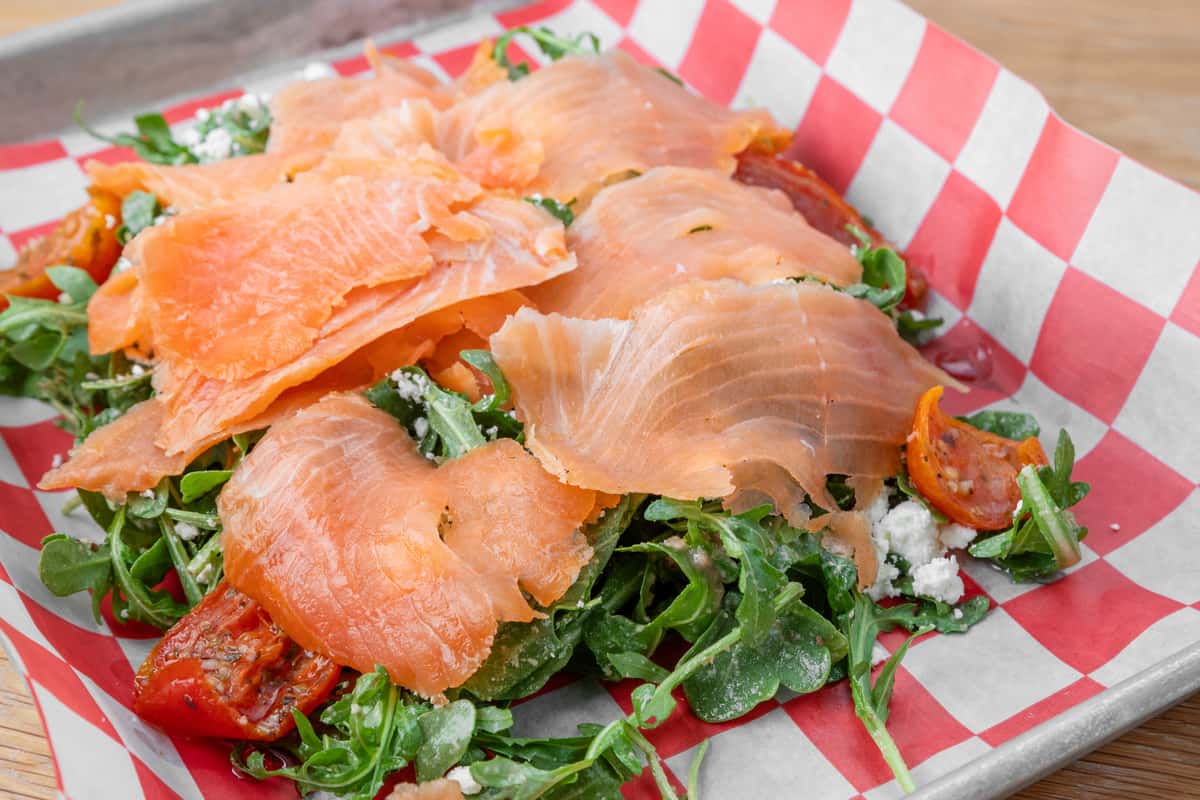 Smoked salmon slices served with fresh greens and salad on red and white checkered paper in a metal tray at a restaurant