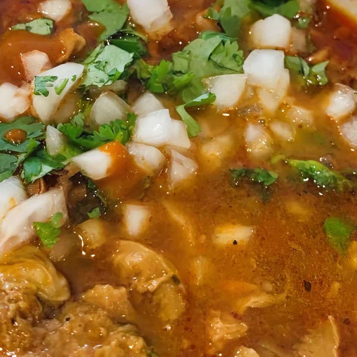 Traditional Mexican menudo soup garnished with chopped onions and fresh cilantro in rich red broth – classic homestyle Mexican soup.