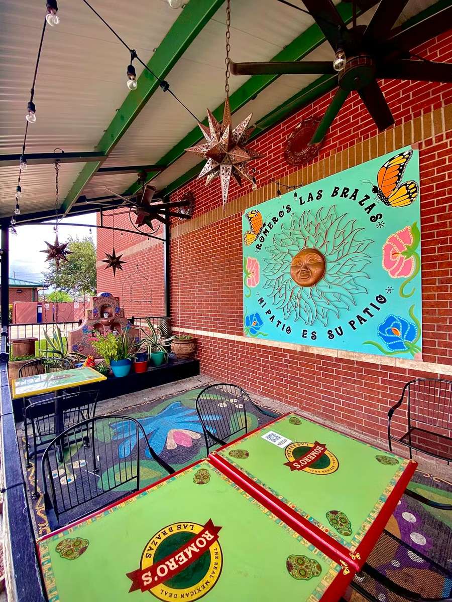 Colorful outdoor patio at Romero's Las Brazas Mexican restaurant featuring bright green tables, decorative metal star lanterns, ceiling fans, and a vibrant mural with a sun and butterflies.