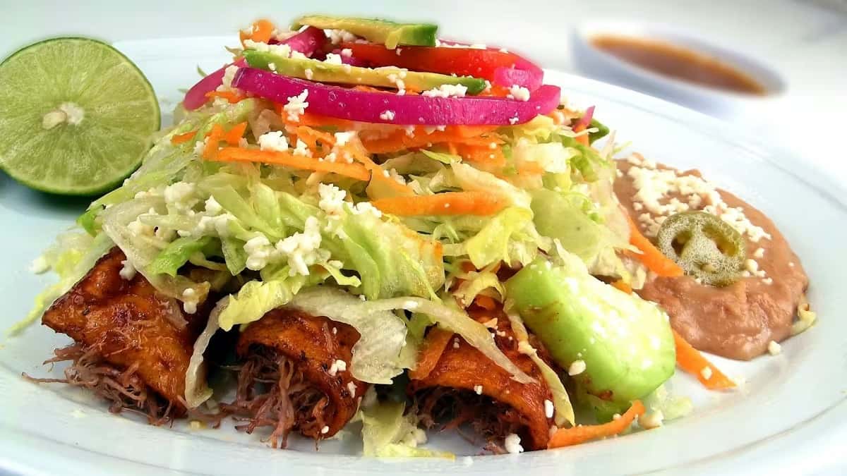 Mexican shredded beef flautas topped with crisp lettuce, pickled red onions, avocado slices, crumbled queso fresco, and shredded carrots, served with refried beans, fresh lime, and salsa on a white plate – vibrant authentic Mexican cuisine presentation.