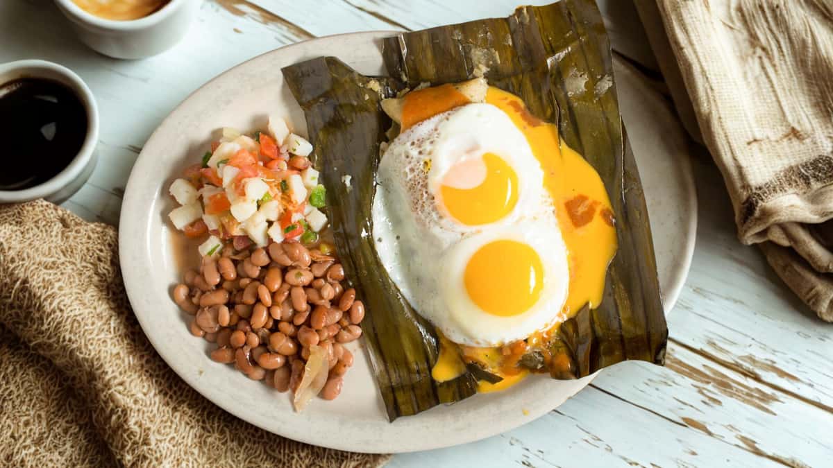 Traditional Mexican breakfast dish served in a banana leaf with two sunny-side-up eggs and melted cheese, accompanied by pinto beans and fresh pico de gallo.
