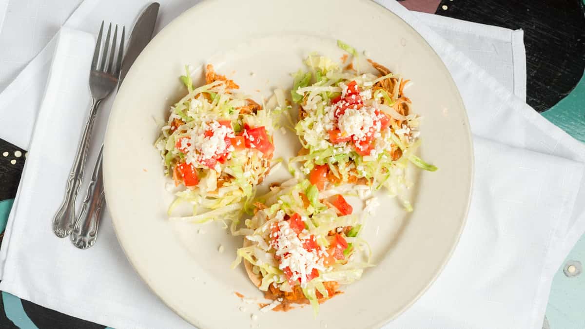 Three crispy shredded chicken tostadas topped with lettuce, diced tomatoes, and queso fresco – classic Mexican appetizer plate.