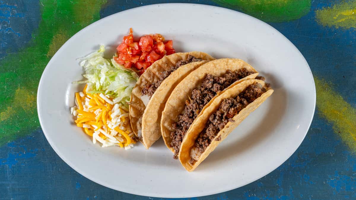 Three ground beef tacos in soft corn tortillas topped with shredded lettuce, diced tomatoes, and blended cheese – authentic Mexican taco platter.