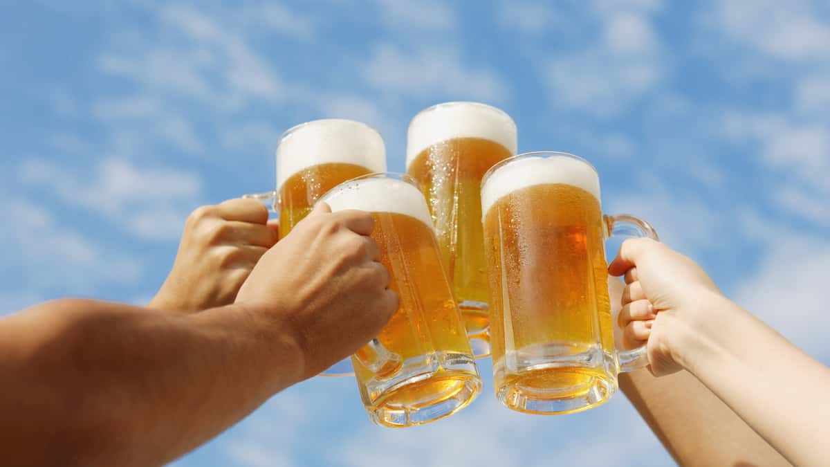 Four people raise and clink frosty glass mugs of golden beer with foamy heads against a bright blue sky.