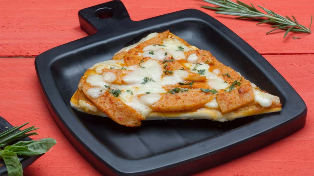 A slice of leftover pizza topped with melted cheese and sliced toppings, served on a black square plate against a red wooden background with sprigs of rosemary and basil nearby.