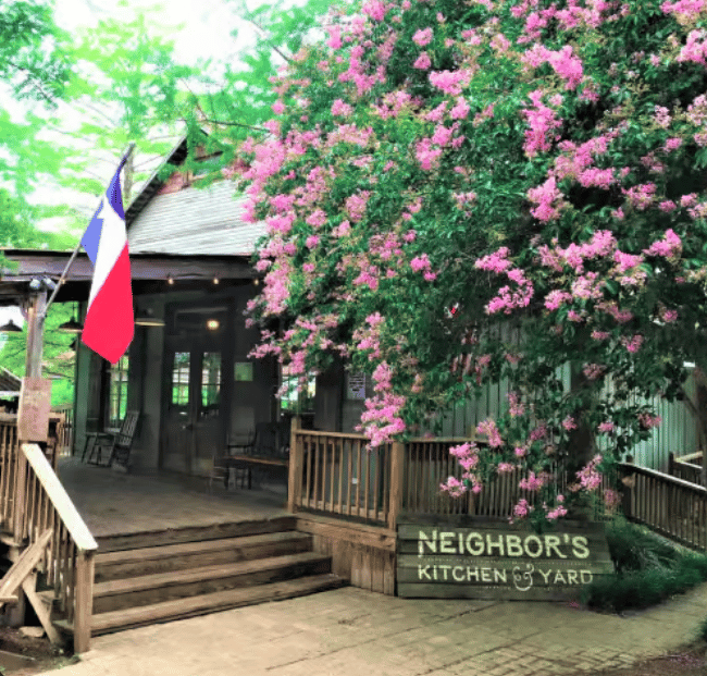 A rustic wooden restaurant entrance with a porch and steps, shaded by a large tree covered in pink blossoms, a Texas flag hanging to the left, and a sign reading