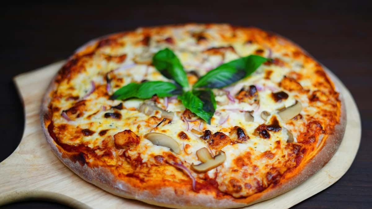 A freshly baked pizza topped with melted cheese, mushrooms, red onions, and basil leaves, served on a wooden pizza board.