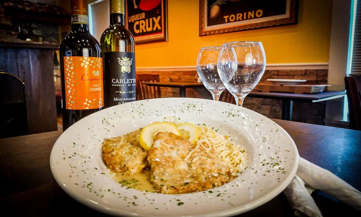 This is a plate of Chicken Piccata served with a side of pasta. The dish features breaded chicken breasts in a lemon butter caper sauce, garnished with lemon slices and sprinkled with herbs and Parmesan. It's elegantly plated with wine and glasses in the background, suggesting a cozy Italian dining setting.