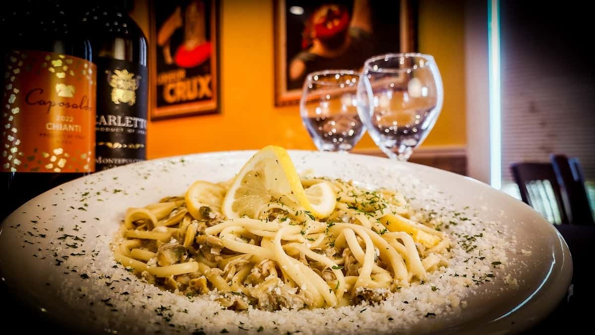 Linguine with clam sauce garnished with lemon slices, surrounded by Parmesan and parsley, wine bottles and glasses in the background.
